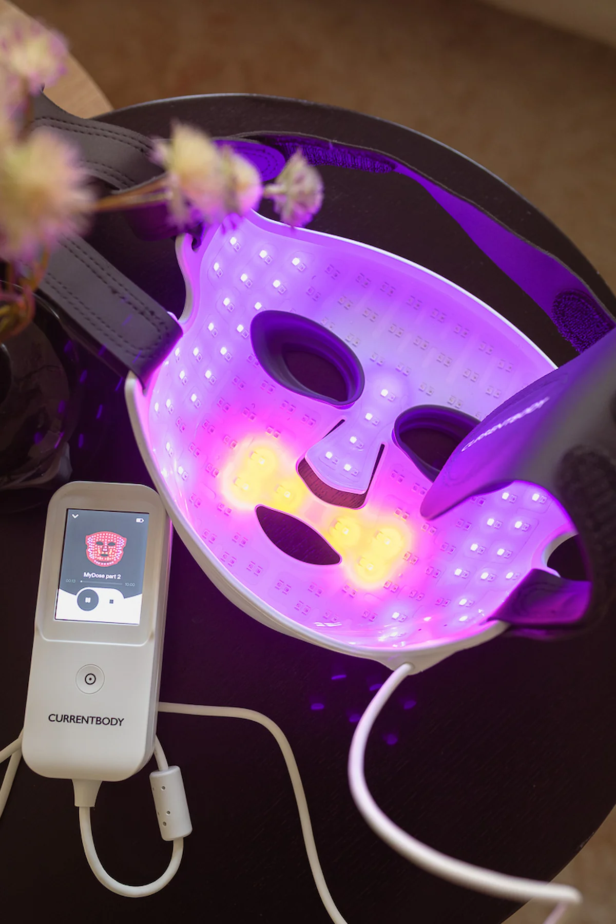 CurrentBody LED-Maske mit mehrfarbig leuchtender Innenseite und Controller auf einem runden Beistelltisch.